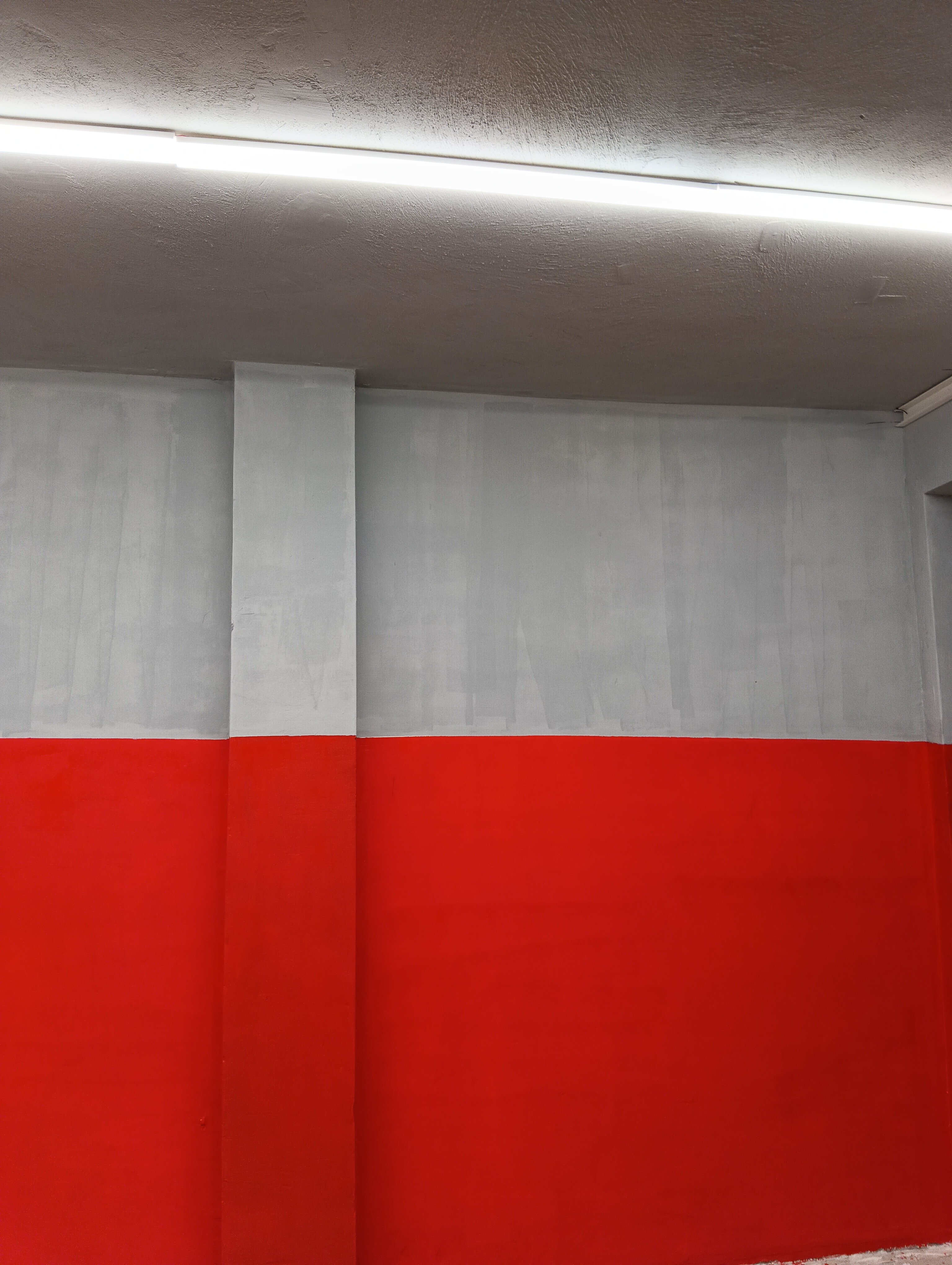 Ένας τοίχος βαμμένος με ένα γκρι πάνω μισό και ένα κόκκινο κάτω μισό, χωρισμένο οριζόντια, με μια κάθετη στήλη και ένα φθορίζον φως στην οροφή.