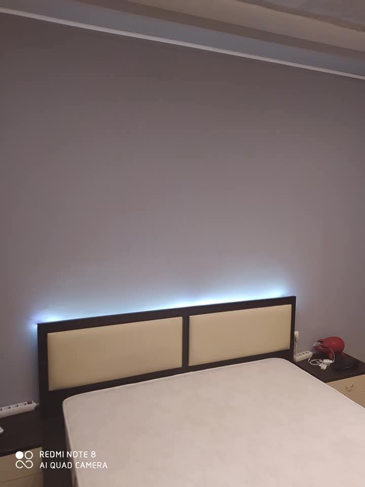 Ένα κρεβάτι με μπεζ κεφαλάρι σε έναν απλό γκρι τοίχο, με μια λωρίδα από μπλε φώτα LED πίσω από το κεφαλάρι και ένα μικρό κομοδίνο με φωτιστικό.